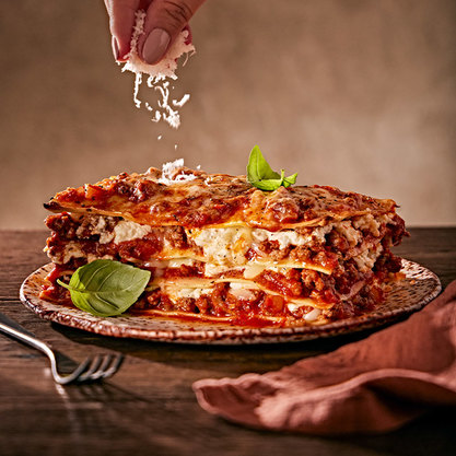 Close-up of a layered lasagna with meat, cheese, and tomato sauce on a plate, topped with fresh basil, while grated cheese is being sprinkled from above.
